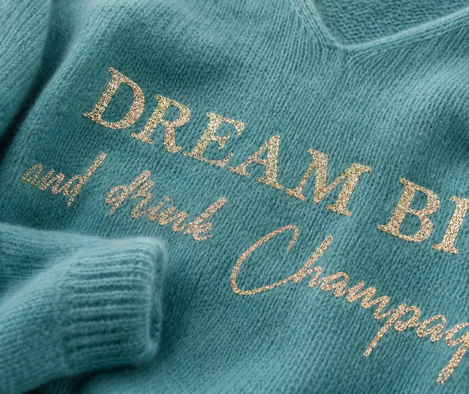 Gros plan du flocage doré pailleté "Dream big and drink Champagne" sur pull céladon col V en Baby alpaga.