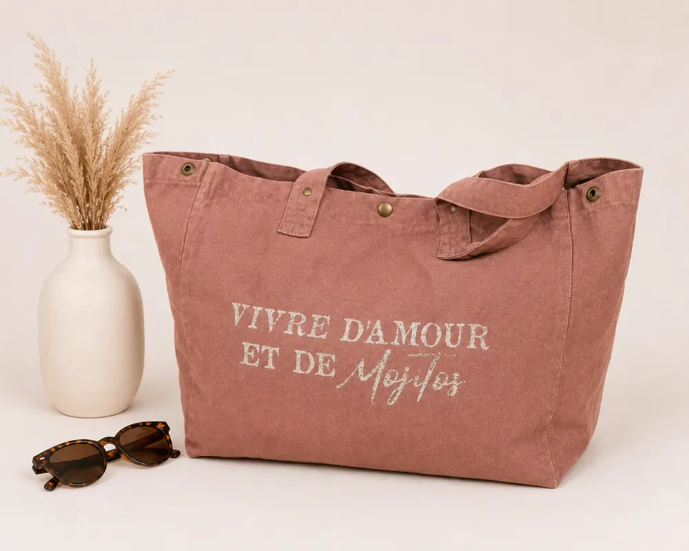Sac cabas santal « Vivre d’amour et de Mojitos » avec flocage doré pailleté, présenté sur un fond clair avec vase, fleurs séchées et lunettes de soleil.
