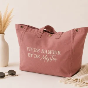 Sac cabas vieux rose « Vivre d’amour et de Mojitos » avec flocage doré pailleté, présenté sur un fond clair avec vase, fleurs séchées et lunettes de soleil.