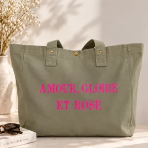 Sac cabas vert sauge « Amour, Gloire et Rosé » avec flocage rose fluo, présenté sur un fond clair et lumineux avec un vase de fleurs séchées, un livre déco et des lunettes de soleil.
