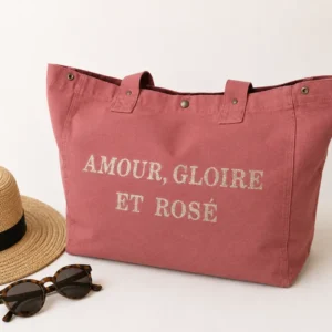 Sac cabas vieux rose « Amour, Gloire et Rosé » avec flocage doré pailleté, présenté sur un fond clair avec un chapeau de paille et des lunettes de soleil.