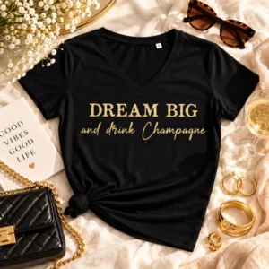 T-shirt femme noir col V « Dream Big and Drink Champagne » en coton, avec flocage doré pailleté, présenté en flatlay sur fond clair avec accessoires élégants : lunettes de soleil, bijoux dorés, sac à main noir et coupe de champagne.
