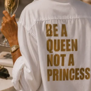 Femme de dos portant la chemise blanche oversize « Be a Queen, not a Princess » floquée doré, une coupe de champagne à la main en terrasse. Création La Strasseuse.