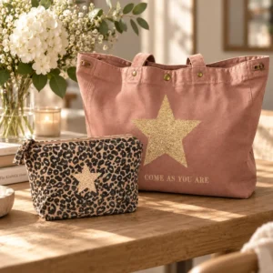 Duo cadeau La Strasseuse : sac cabas XXL santal « Come As You Are » à grande étoile dorée pailletée et trousse de toilette léopard à étoile champagne, posés sur une table en bois près d'un bouquet de fleurs blanches.