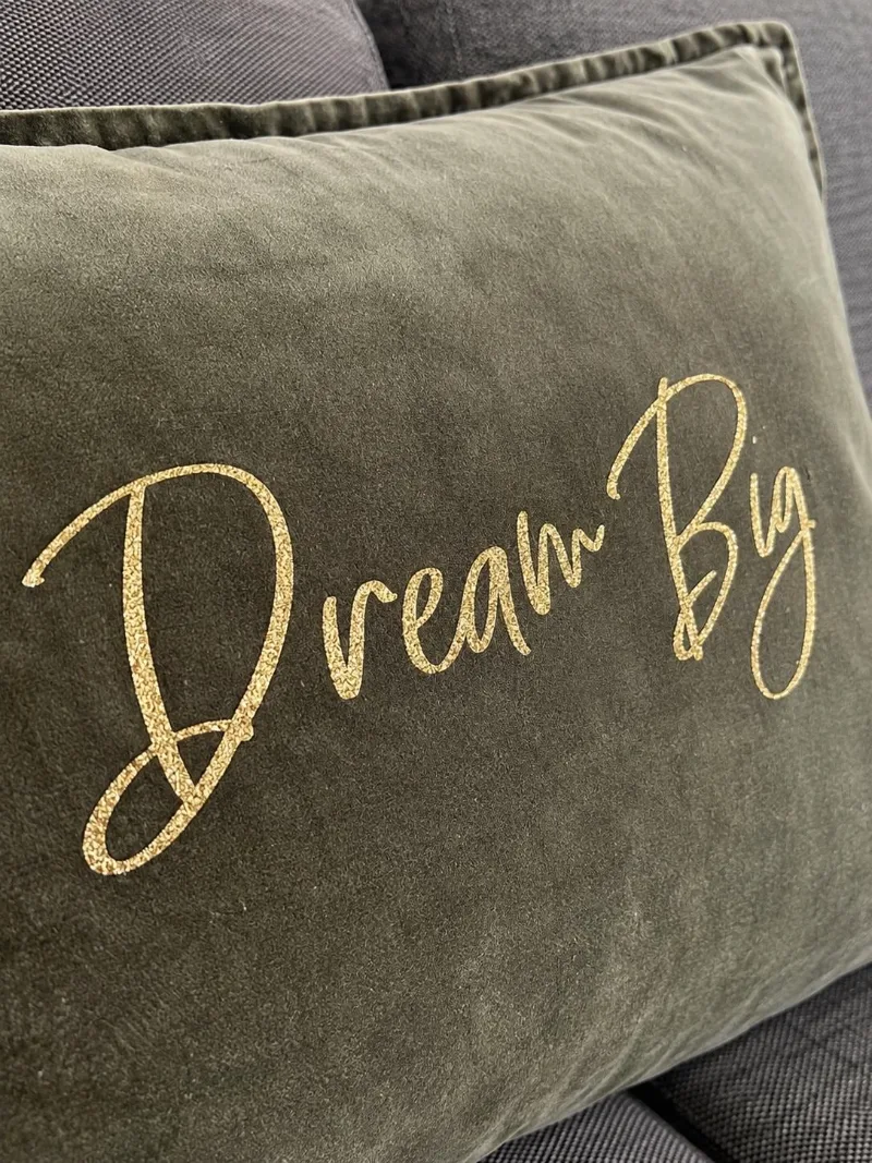 Gros plan sur la housse de coussin en velours de coton kaki et son slogan Dream Big en écriture manuscrite dorée pailletée qui scintille à la lumière.