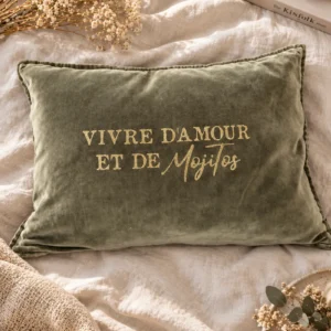 Vue du dessus de la housse de coussin rectangulaire en velours de coton kaki avec le slogan Vivre d'Amour et de Mojitos en lettres dorées pailletées, posée sur un drap en lin crème avec des fleurs séchées, un livre et une bougie.