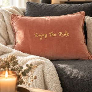 Housse de coussin rectangulaire en velours de coton vieux rose avec le slogan Enjoy the Ride en écriture manuscrite dorée, posée sur un canapé gris anthracite avec un plaid blanc moelleux et une bougie allumée.
