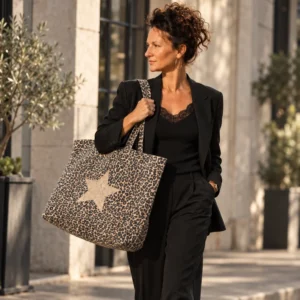 Femme en tailleur noir et caraco en dentelle portant à l'épaule le maxi tote bag léopard à grande étoile champagne pailletée devant une façade en pierre