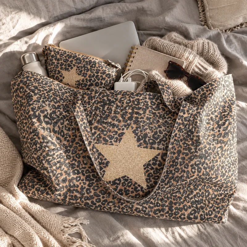 Maxi tote bag léopard étoile champagne rempli d'un ordinateur, d'une gourde, d'une trousse léopard étoile assortie, d'un carnet, de lunettes et d'un pull, posé sur un lit