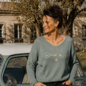 Femme portant le pull col V kaki clair en baby alpaga « Strasser sa route » en doré pailleté avec son étoile, et un jean brut, devant une voiture vintage. Création La Strasseuse.