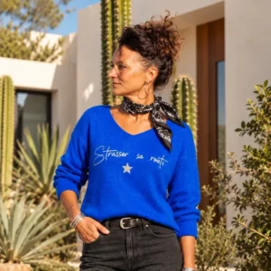 Femme portant le pull col V bleu roi « Strasser sa route » en argent pailleté avec son étoile, un foulard noir et un jean noir, dans un jardin méditerranéen. Création La Strasseuse.