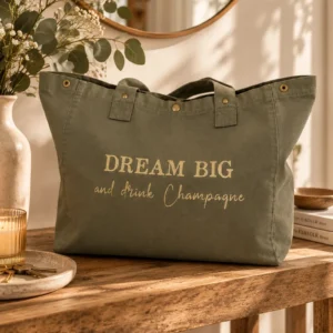 Sac fourre-tout kaki « Dream Big and drink Champagne » en or pailleté posé sur une console en bois, devant un miroir. Création La Strasseuse.