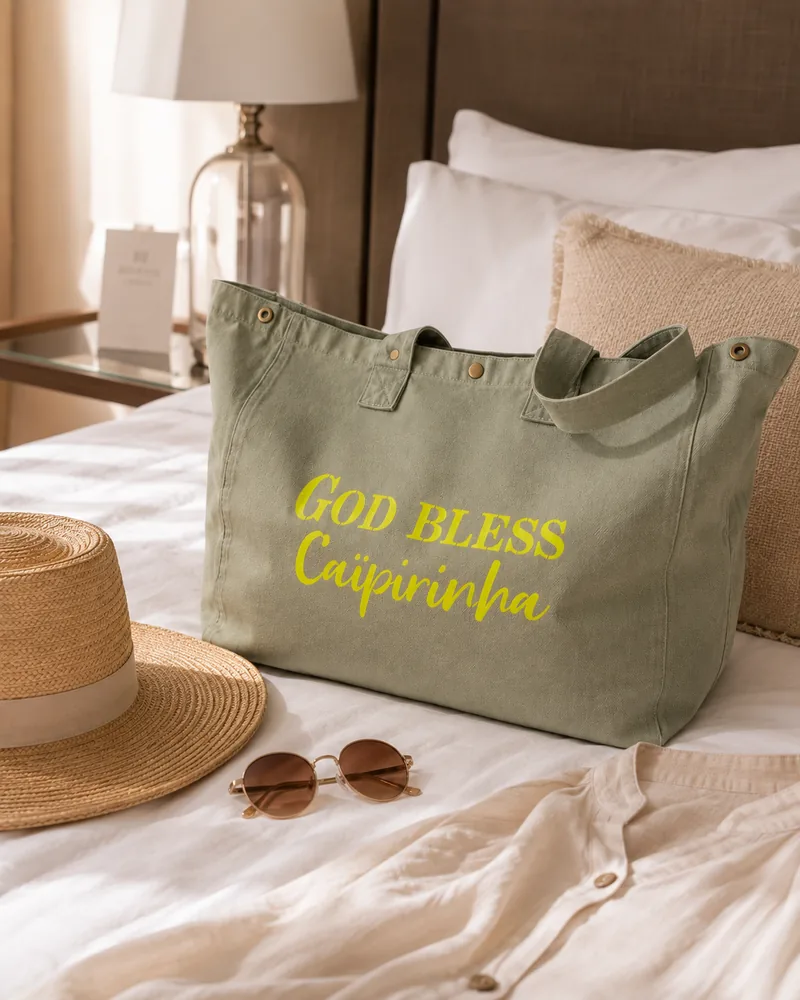 Sac fourre-tout vert sauge "God bless Caïpirinha" en jaune fluo, posé sur un lit avec chapeau de paille et lunettes de soleil. Création La Strasseuse.