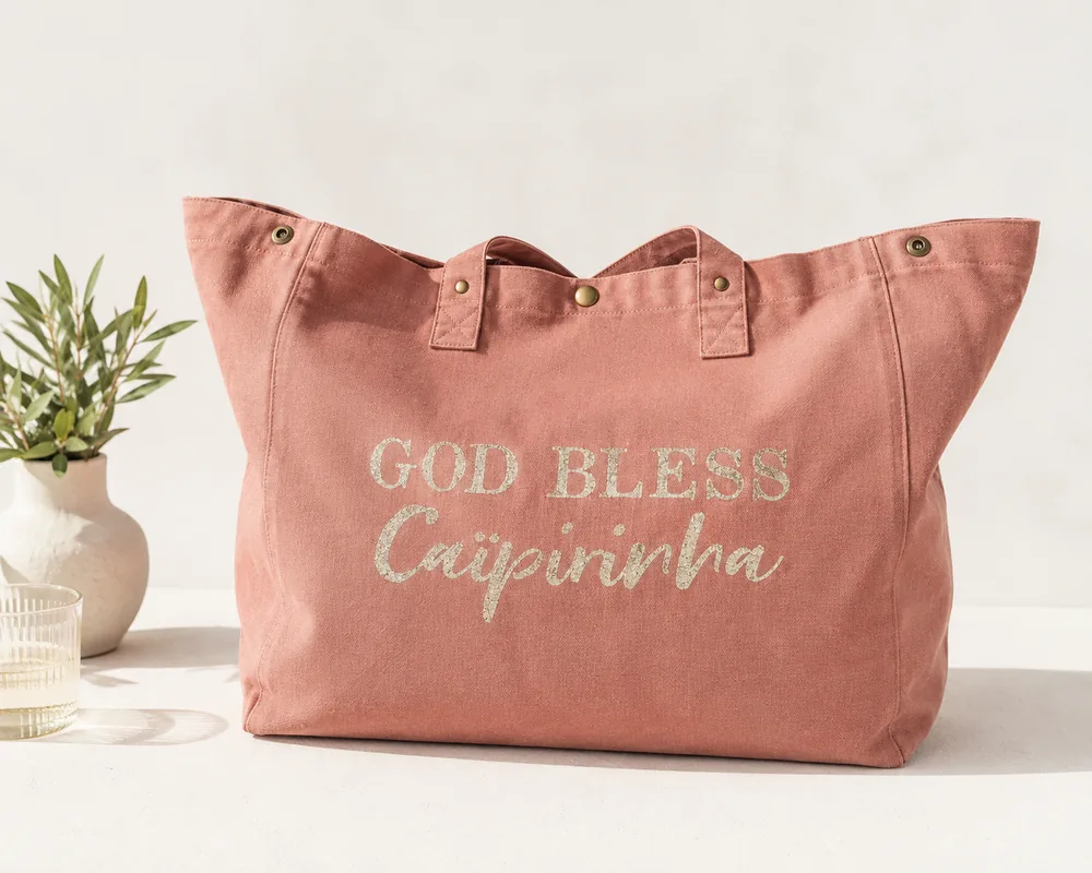 Sac fourre-tout vieux rose avec "God bless Caïpirinha" en or pailleté, sur une table claire avec une branche d'olivier. Création La Strasseuse.