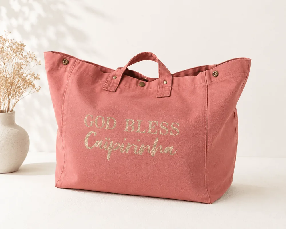 Sac fourre-tout vieux rose avec "God bless Caïpirinha" en or pailleté, sur fond clair à côté d'un vase de fleurs séchées. Création La Strasseuse.