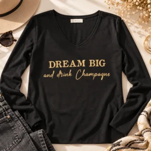 Top manches longues noir col V « Dream Big and drink Champagne » en or pailleté, présenté à plat avec un jean gris, un chapeau et des bijoux dorés. Création La Strasseuse.