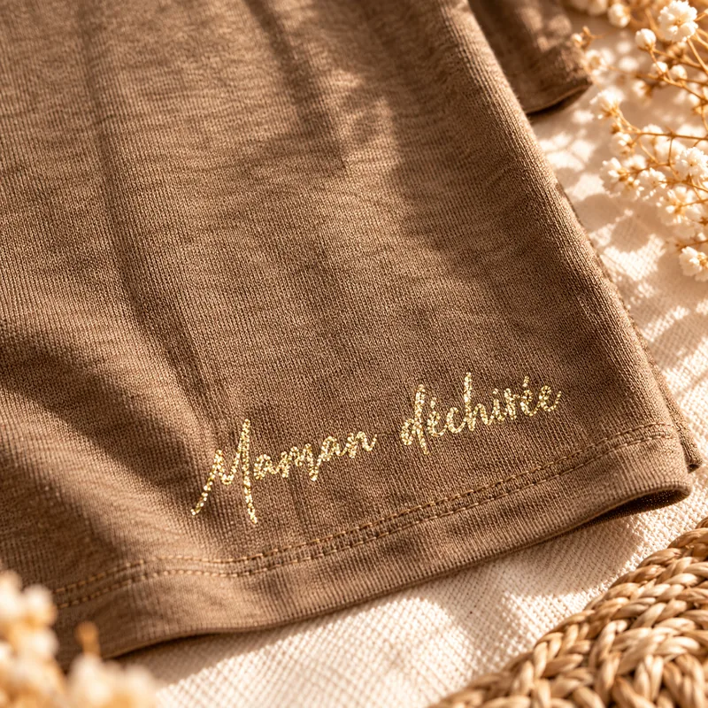 Détail de l'inscription « Maman déchirée » en or pailleté, en petit au bas du dos du top manches longues. Création La Strasseuse.