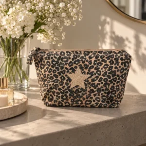 Trousse de toilette léopard avec étoile champagne pailletée posée sur une console près d'un bouquet de fleurs blanches et d'un parfum