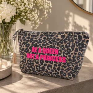 Trousse de toilette léopard au flocage rose fluo « Be a Queen, Not a Princess », posée sur une console en pierre devant un bouquet de gypsophile et un miroir doré.