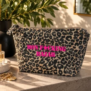Trousse de toilette léopard au flocage rose fluo « Mon F*cking Bazar », posée sur une console en pierre à côté d'une plante verte, d'un parfum et d'une chaîne dorée.